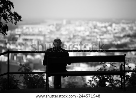 Senior is sitting alone on the bench looking at city at evening. A lonely old man sitting on a bench in a park.Elderly gentleman on park bench in contemplation,black and white photo