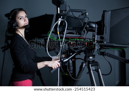 Recording at TV studio.Professional camera operator with headphones and camera in television news broadcast.Camerawoman working with big broadcasting camera.Positioned video camera.Behind the scenes