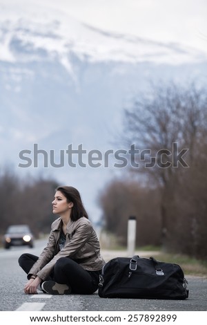 Young woman hitchhiking on countryside road.Traveler woman sitting alone along the road.Pretty young woman tourist hitchhiking.Left alone on the road and lost