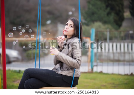 Beautiful woman blowing bubbles on the playground.Young woman blowing bubbles swinging in the park.Gorgeous flirty brunette woman starts soap bubbles in park