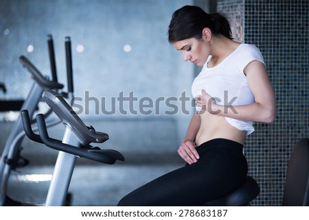 Woman riding an exercise bike in gym.Cardio and fat loss workout.Sport and fitness,summer body goals.Fit sexy woman holding showing stomach area.Healthy nutrition,fat lose,healthy lifestyle  concept.