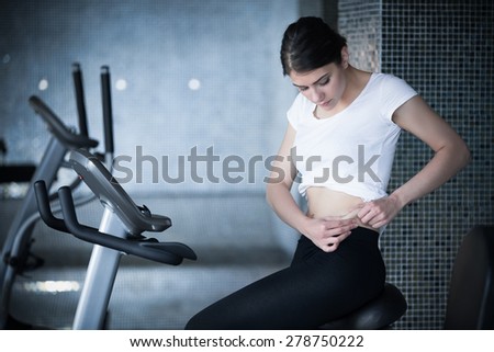 Woman riding an exercise bike in gym.Cardio and fat loss workout.Sport and fitness,summer body goals.Fit sexy woman pinching showing stomach area.Healthy nutrition,fat lose,healthy lifestyle  concept.