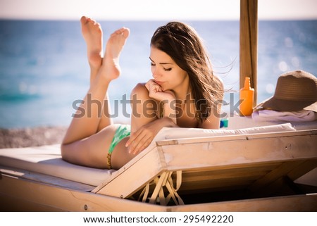 Smiling beautiful woman sunbathing in a bikini on a beach at tropical travel resort, enjoying summer holidays.Young woman lying on sun lounger near the sea.Happy serene woman having SPF protection