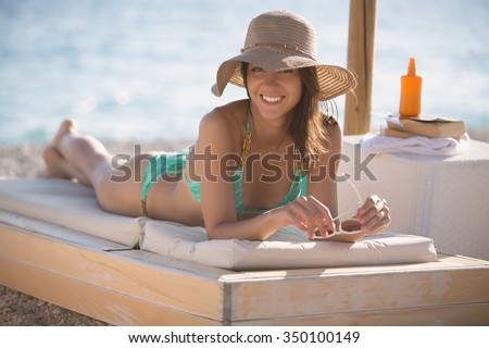 Smiling beautiful woman sunbathing in a bikini on a beach at tropical travel resort, enjoying summer holidays. Young woman lying on sun lounger near the sea. Happy serene woman having SPF protection