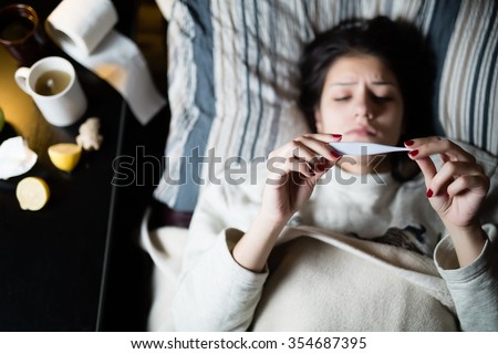 Sick young woman in bed at home having flu,measuring temperature.Thermometer to check temperature for fever.Flu.Virus.Sick woman laying in bed.Focus on thermometer