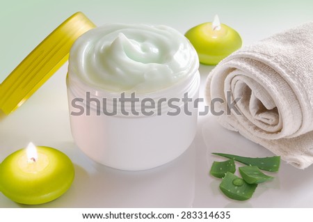 White plastic container with facial or body cream of aloe vera. Candles, towel and plant decoration and green background isolated. Top view