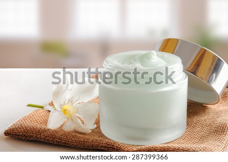 Glass open jar with facial or body cream on burlap. With lid and flower.Windows background. Front view.