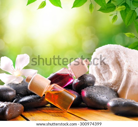 Oil and essences for body care and towel with black stones on wood base. Green leaves and bokeh background. Square composition.