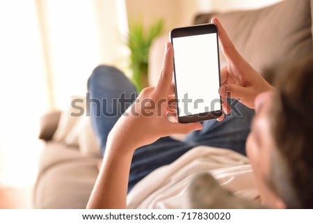 young man watching multimedia content on a smartphone vertical lying on a couch in the living room at home