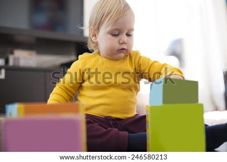 blonde sixteen month baby yellow sweater playing with color boxes inside home
