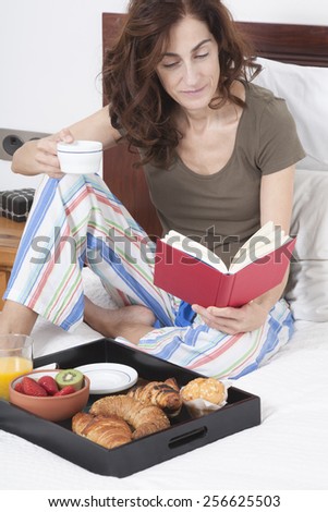 brunette woman green shirt striped pajama pants sitting on white bed reading red book with breakfast tray croissants orange juice strawberry kiwi cupcake red rose flower