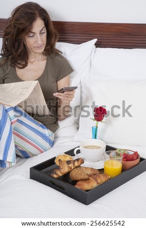woman green shirt striped pajama pants sitting on white bed reading financial newspaper and phone with breakfast tray croissants orange juice strawberry kiwi cupcake red rose flower
