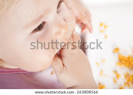 overhead shot of one year age caucasian baby pink plastic bib eating meal yellow orange rice paella with her hand in white high-chair