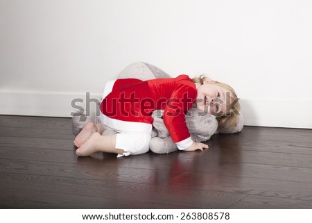one year age caucasian blonde cute lovely tender baby barefoot Santa Claus Christmas holidays disguise lying embraced over grey plush doll on brown wooden floor white wall