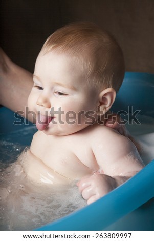 six months age blonde baby body and face sticking out tongue washing by woman mother hands in blue little plastic bath indoor with brown background