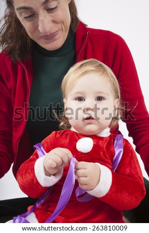 one year age caucasian blonde baby Santa Claus disguise with brunette woman mother red cardigan green trousers opening silver wrapped paper box gift Christmas on white background
