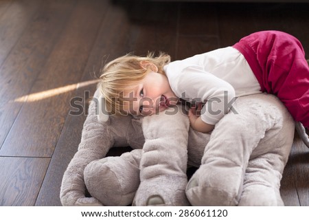 portrait of happy blonde caucasian baby nineteen month age looking at camera smiling  face embraced grey plush doll on brown wooden floor