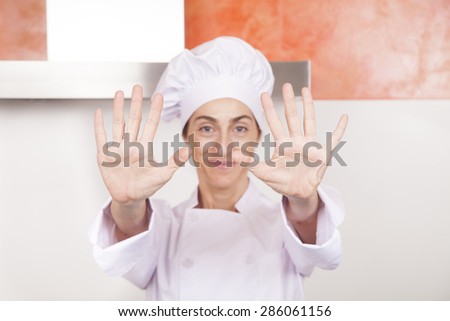 portrait of brunette happy chef woman with professional jacket and hat in white and orange kitchen showing both open palms of hands