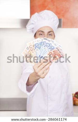 wad of fifty twenty and ten Euro banknotes wad in hands of brunette happy chef woman with professional jacket and hat in white and orange kitchen