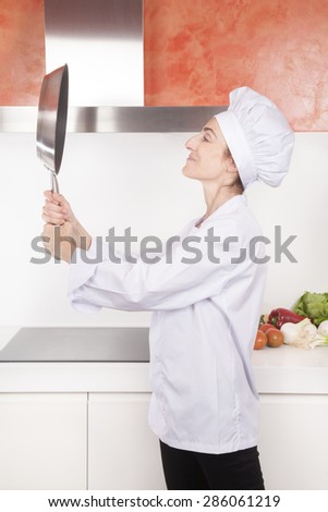 portrait of happy brunette chef woman with professional jacket and hat in white and orange kitchen looking herself at steel blank pan like a mirror