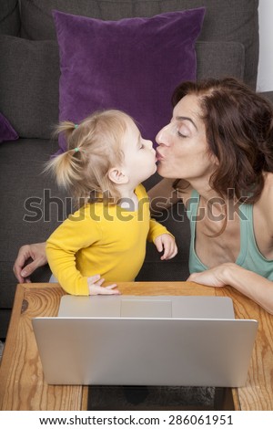 nineteen month aged blonde baby yellow shirt with pigtails and brunette woman mother kissing in mouth with grey laptop on brown table