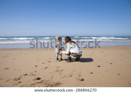 two years age blonde baby with grey coat and brunette mother woman squatting in sand of beach in front of water sea or ocean