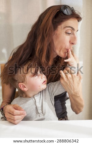 brunette woman mother with sunglasses on head sucking finger of her blonde baby two years age with grey shirt chocolate smeared face sitting in white table