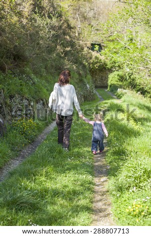 two years age blonde baby with blue dress pigtails and brunette mother woman with grey jersey back walking holding hand in green path in mountain