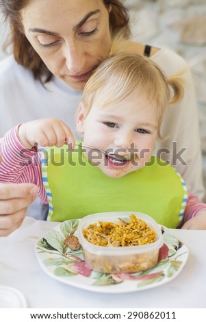 portrait of two years age blonde baby with green bib eating rice paella from tupperware sitting on legs of mother woman in white table