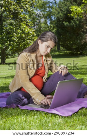 pregnant young woman with orange shirt touching laptop sitting at a green park in Madrid Spain Europe