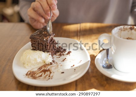 woman hands taking chocolate cake piece with spoon and white small cup cappuccino coffee on light brown wooden table