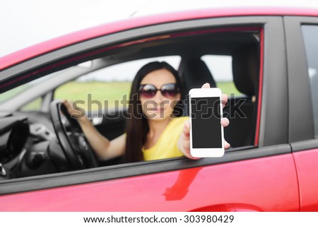 Woman showing her smartphone with empty screen out the window of a car. Focus on model.