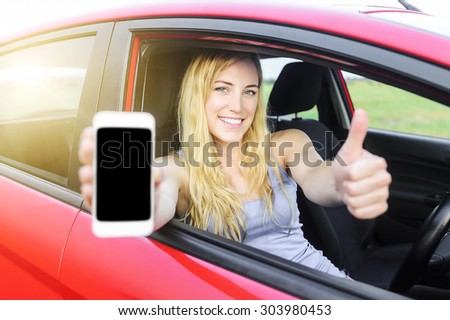 Happy blonde woman showing smartphone and showing thumb up out the window of a car. Focus on model.