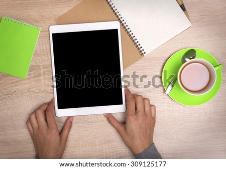 Tablet PC in the hands, a cup of tea and notebook on the desktop. Top view.