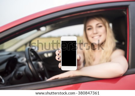 Happy blonde woman showing smartphone out the window of a car. Focus on mobile phone.