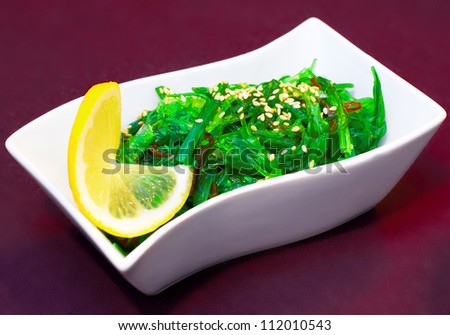Japanese salad seaweed green with onion and vegetables with a slice of lemon on a white plate on a dark red background