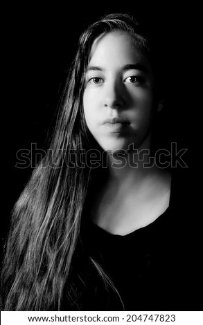 Dramatic low-key Rembrandt lighting portrait of an 18 year old young woman with long hair on black background in black and white (B&W)