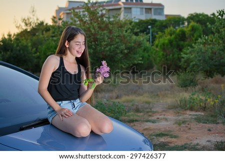 A 13 year old teenage girl with braces on her teeth sitting on a car enjoying pink flowers and the summer sunset