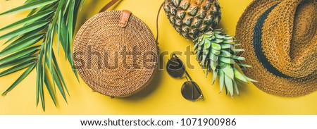 Colorful summer female fashion outfit flat-lay. Straw hat, bamboo bag, sunglasses, palm branches, pineapple over yellow background, top view, wide composition. Summer fashion, holiday concept