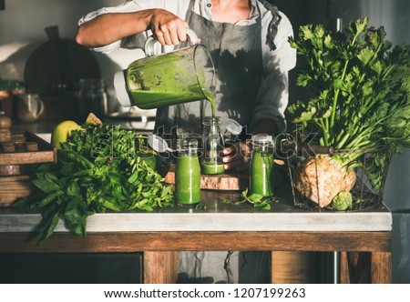 Making green detox take-away smoothie. Woman in linen apron pouring green smoothie drink from blender to bottle surrounded with vegetables and greens. Healthy, weight loss food concept