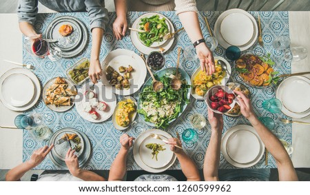 Mediterranean style dinner. Flat-lay of table with salads, starters, pastries over blue table cloth with hands holding drinks, sharing food, top view. Holiday gathering and vegetarian party concept