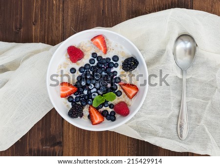 A bowl of oat porridge with fresh berries, honey, almond petals and leaves of mint over a piece beige linen fabric on a dark wooden background, a view from above