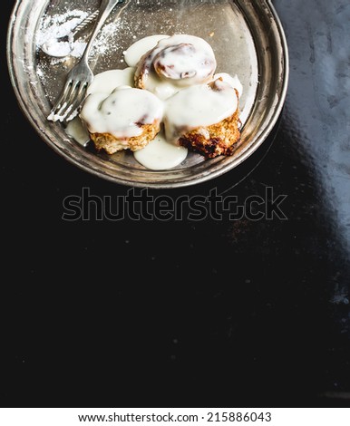 Cinnamon rolls with cream icing and sugar powder on a metal dish with fork a tea-spoon over a black metal surface