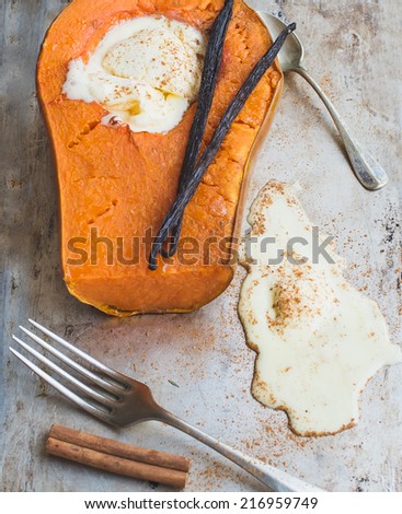 Backed pumpkin half with a scoop of vanilla ice-cream, vanilla sticks, cinnamon, a fork and a tea-spoon over a rough metal surface