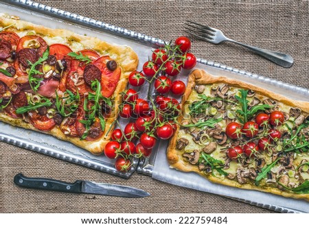 Two rustic square pizzas with fresh arugula and cherry-tomatoes on silver trays over a sackcloth background
