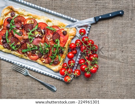 Homemade square pizza with meat, salami, cherry-tomatoes and fresh arugula on a silver tray over a sackcloth background