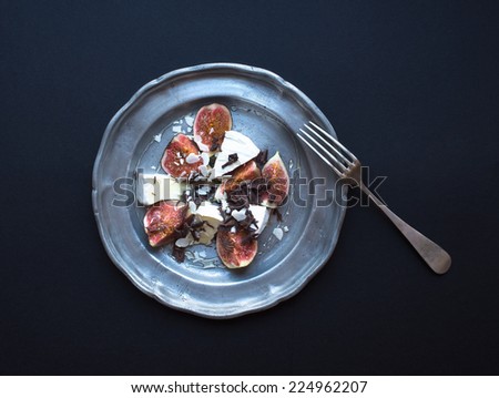 Dessert consisting of fresh figs and brie cheese with honey, almond petals and chocolate chips on a metal plate over a dark background