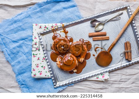 Cinnamon rolls with warm caramel topping and cinnamon sticks on a silver tray with tea-spoons over pieces of colorful fabric