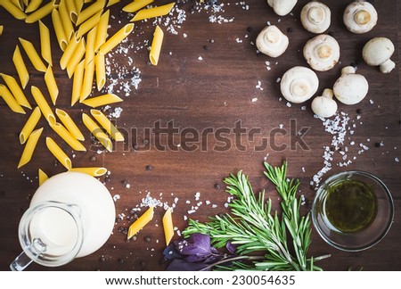 Mushroom pasta ingredients: penne, mushrooms, a jug of cream, pesto sauce, fresh herbs and spices on a dark wood background with a copy space in the center