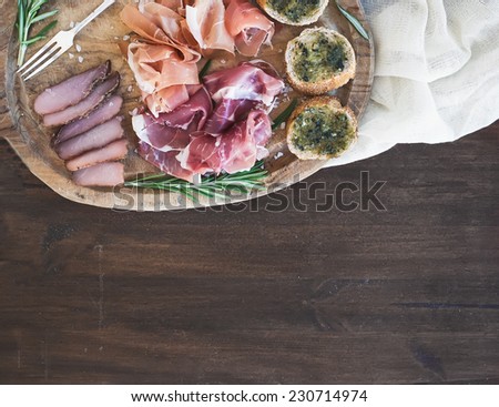 Wine meat appetizer set: prosciutto, serrano and cured lamb meat selection with spices, herbs and baguette slices with pesto on a rustic wooden board over a dark wood background with a copy space